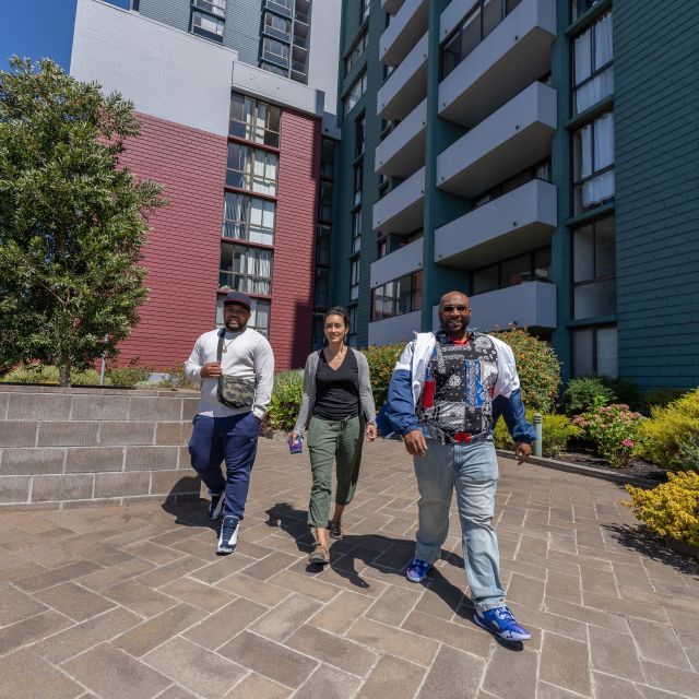 Three people walking forward with a tall apartment complex in the background. 