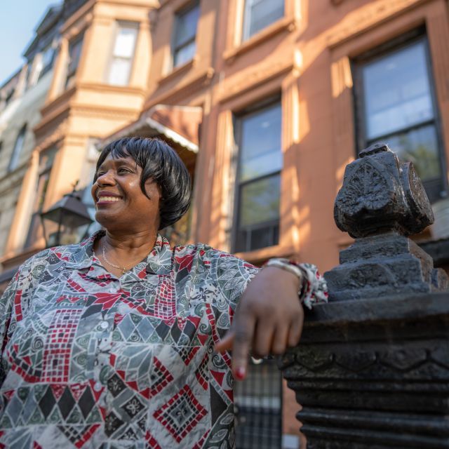 Woman stands on a sidewalk leaning on the banister of the brownstone steps