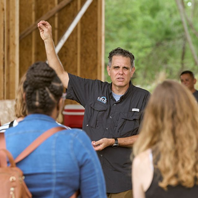 A man standing outside talking to a group of people while gesturing to what is behind him