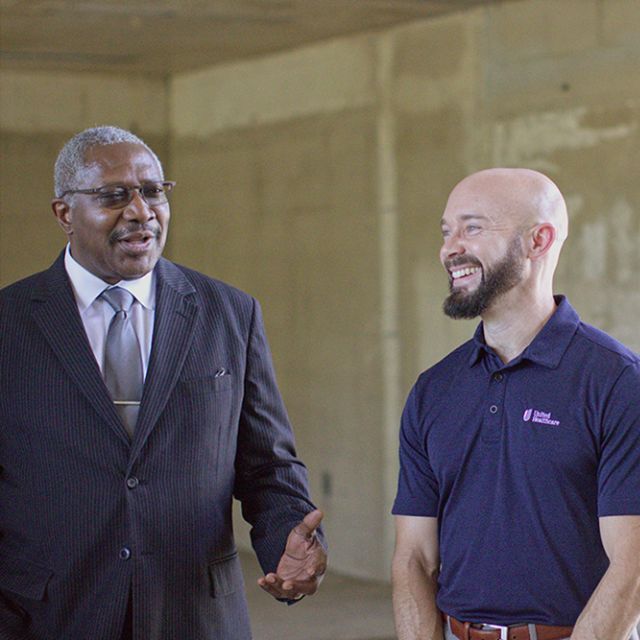 One man in a navy blue shirt smiling while a man in a suit talks to him