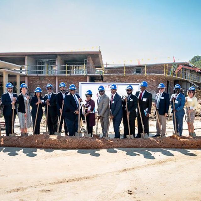 A line of people with blue hard hats on stand holding shovels at a groundbreaking ceremony