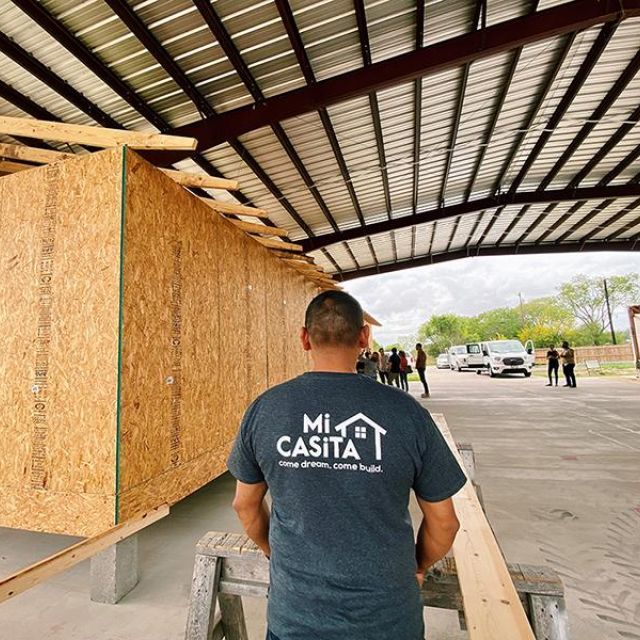 A man stands in front of a home under construction. He is wearing a shirt that has "MiCASiTA, come dream. come build." printed on it.