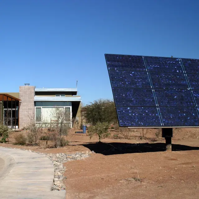 Desert setting with large ground-mounted solar panel in the forefront and a low-rise adobe-style apartment community in the background.