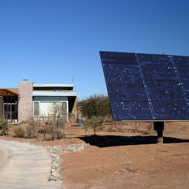 Desert setting with large ground-mounted solar panel in the forefront and a low-rise adobe-style apartment community in the background.