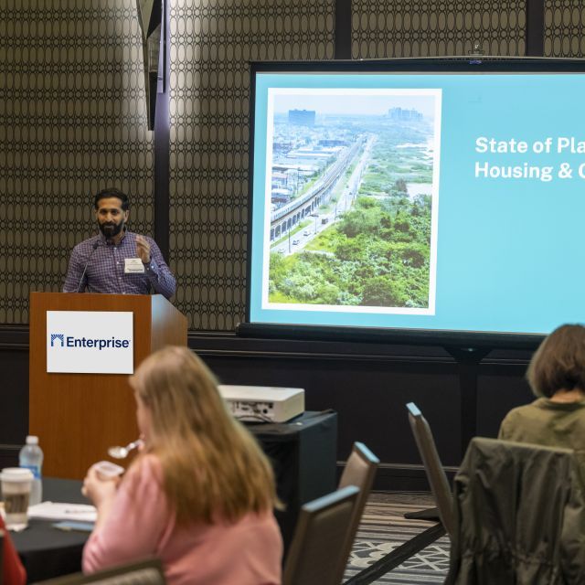 Speaker stands at a podium speaking to an audience in a conference room. To the speaker's right is a screen with a photo showing an aerial view of a city and the text: State of Play: Housing & Climage