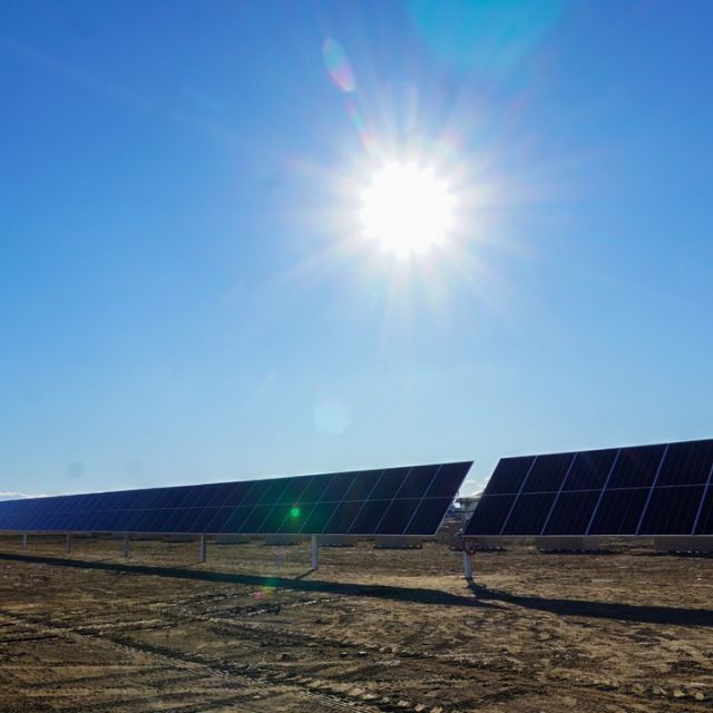 A long row of solar panels with brown open space in the background and a bright white sun shining overhead.