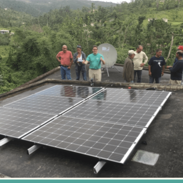 Group of people stand before a flat solar panel with green trees and hills in the background.