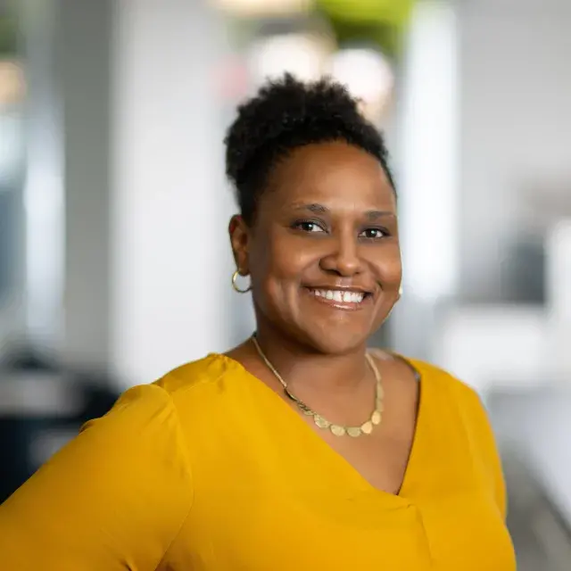 Person smiling, wearing their hair in bun and a golden yellow blouse
