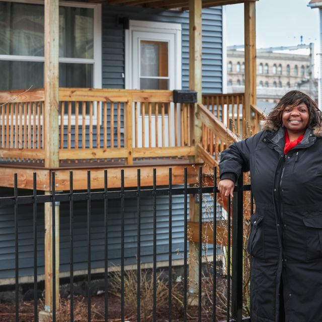 Person with shoulder-length brown hair wearing long black parka stands in front of a blue house with a front porch; a building with a crane on top is in the background.