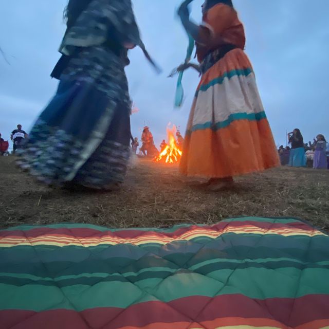 People wearing colorful skirts dance on a field with a bonfire burning in the background and a colorful blanket in the foreground