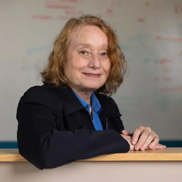 Person with shoulder length red hair wearing a black blazer and blue blouse and a whiteboard in the background