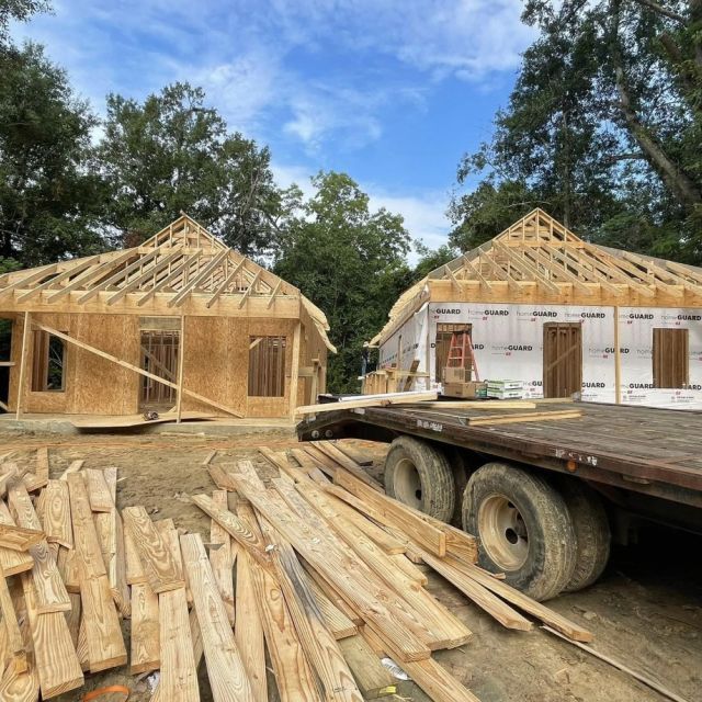 Two homes under construction stand side by side with a blue sky and treetops in the background