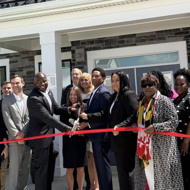 Smiling people in business attire standing in front of a building and cutting a red ribbon.