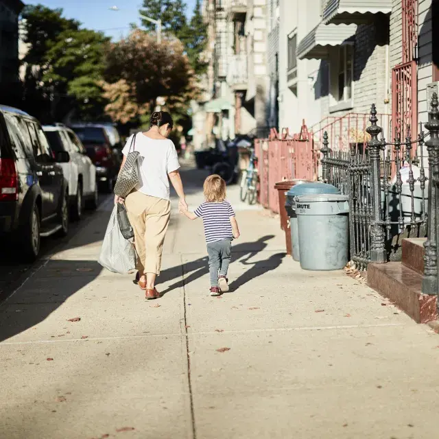 Mother and child walk on a neighborhood sidewalk along apartments, homes, and parked cars