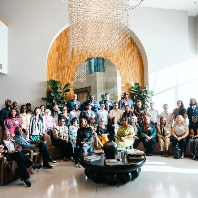 A group of people pose together in a sunlit room with high ceiling in front of a white wall with a tan wooden arch panel