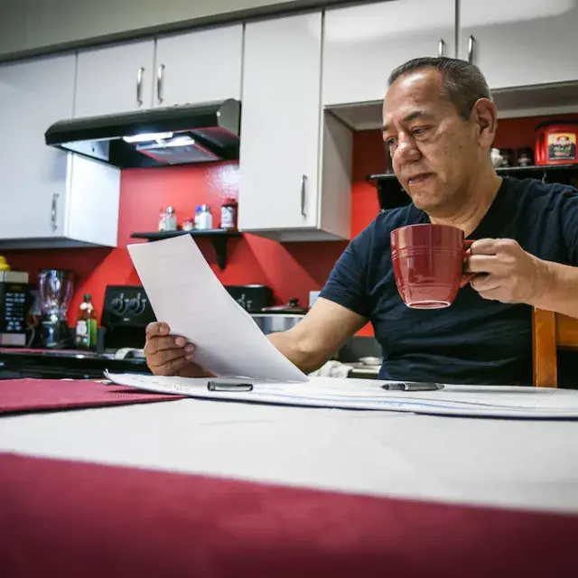 Man in dark shirt sitting at kitchen table drinking coffee and looking at paperwork