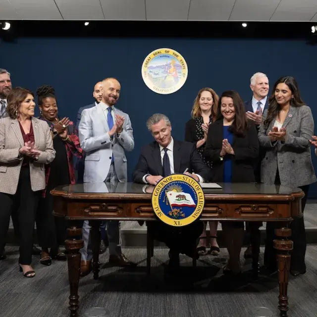 California Gov. Gavin Newsom sitting at desk signing a document, surrounded by clapping people