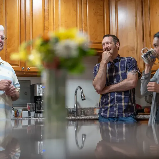 Three people stand in a kitchen smiling and talking with a vase with flowers in the center.