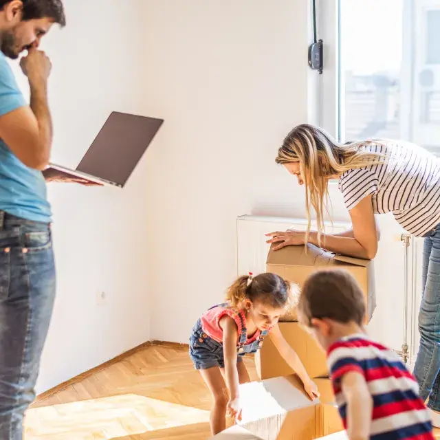 Family stands in sunlit room moving boxes while person stands staring at a laptop screen and looking uneasy