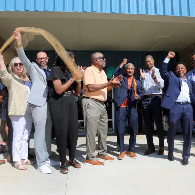 A group of people stand smiling and holding a ribbon in front of a building entrance