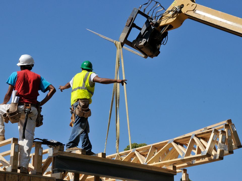 Two construction workers standing on beams as additional pieces are lowered by a crane