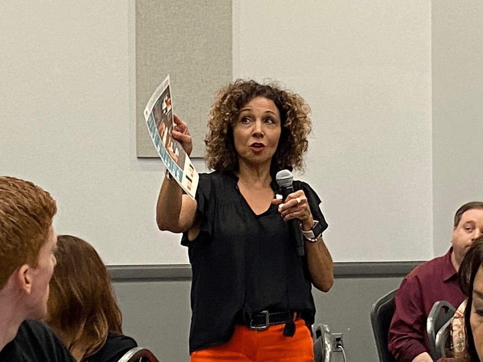 A woman standing and speaking to a group while pointing with a piece of paper in her hand
