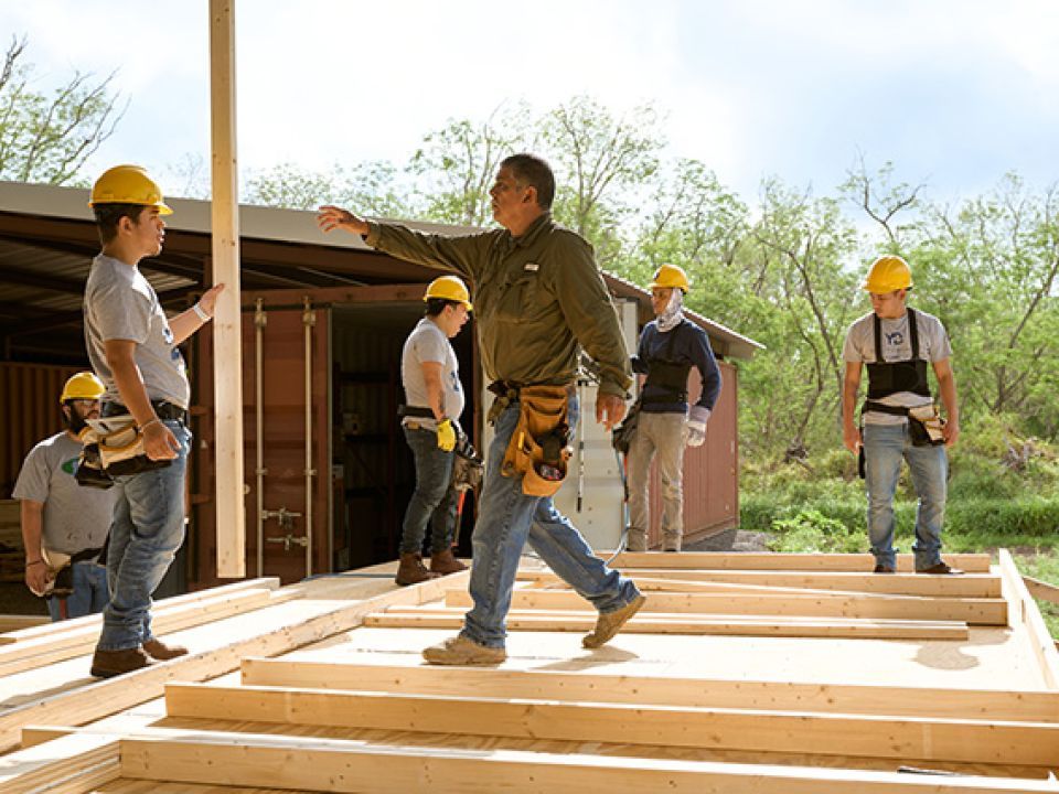 A crew of workers on a job site. A worker reaches out for a piece of lumber another worker is holding