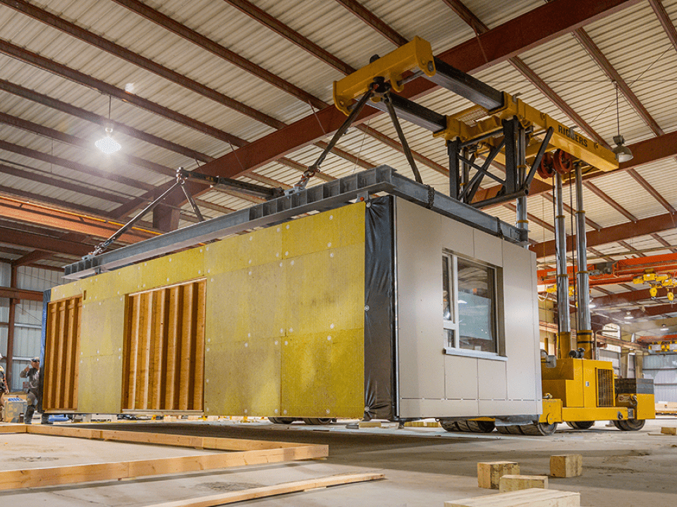 Two construction workers examine a pre-built room lifted by specialized lifting equipment in a warehouse
