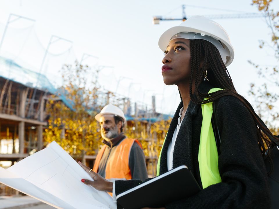 A construction worker holds building plans at a site as she looks skyward. Another construction worker stands nearby
