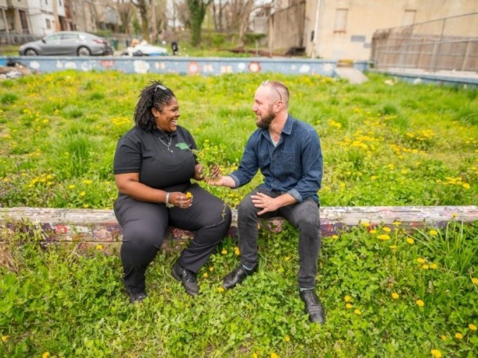 Two people sit and talk on a stone wall in a grassy area of a park