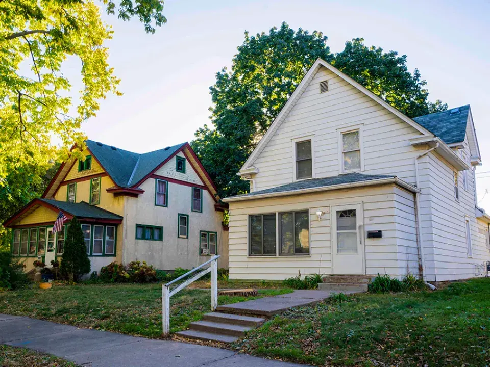 Two detached single-family homes on a residential street, surrounded by mature trees and lawns, with a sidewalk and front steps leading to the houses.