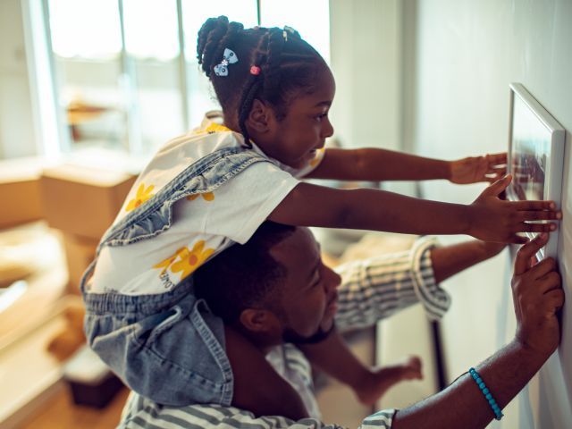 Girl sitting on her father's shoulders helps hang a picture in their home