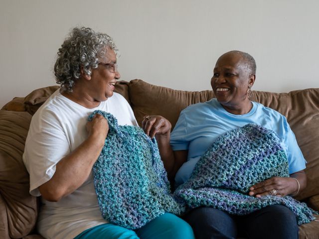 Two smiling women on couch holding up their colorful, blue crochet squares.