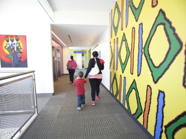 Children and adults walk down a hallway by a yellow wall painted with multi-colored shapes