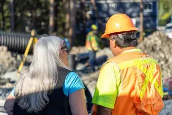 A man in an orange hard hat and vest speaking to a woman on a job site