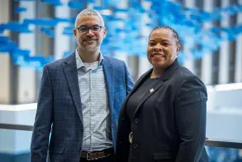 Two people stand together in a room with artwork in blue behind them